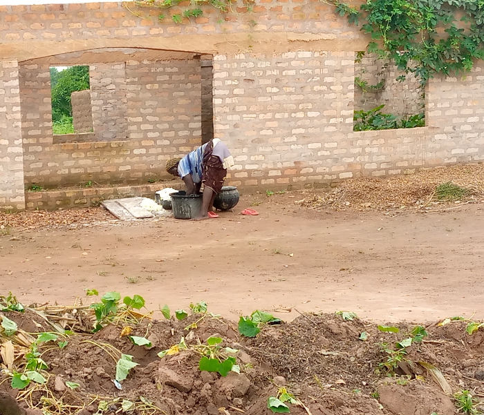 Person fetching water from a well outside a brick building in a rural setting.