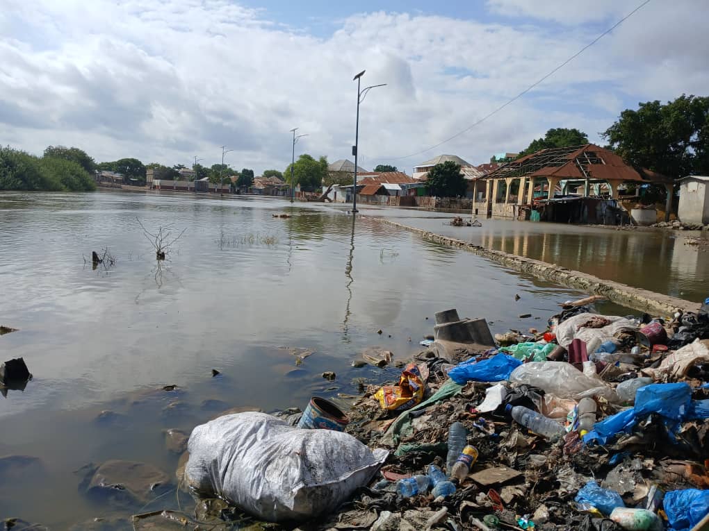 Flooded street with debris and trash piled in the foreground and partially submerged buildings in the background under a cloudy sky.