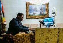 Man in a suit podcasting with a mic and laptop in a room with a classic painting and a ring light.