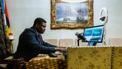 Man in a suit podcasting with a mic and laptop in a room with a classic painting and a ring light.