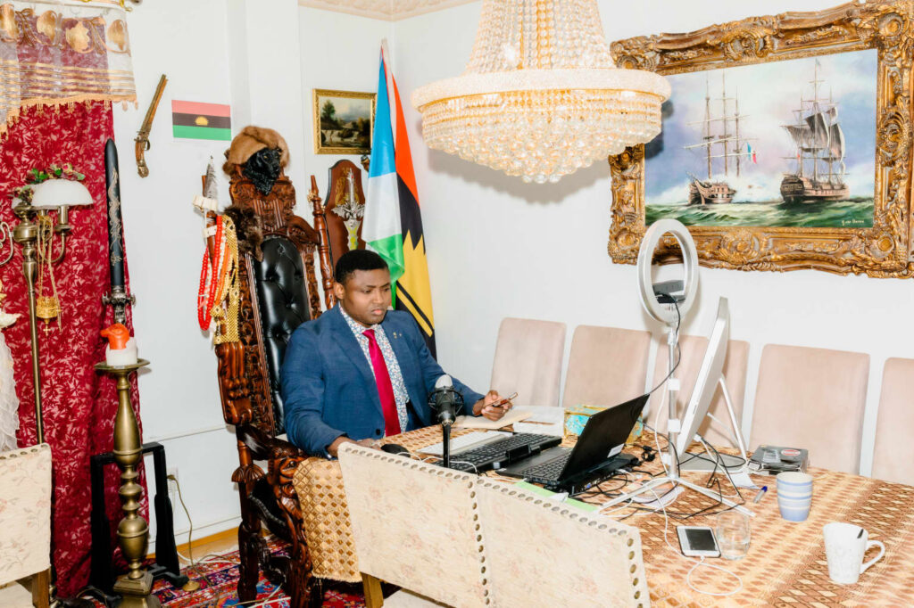 A man in a suit works at a desk amid eclectic decor with a flag, traditional objects, and artwork.