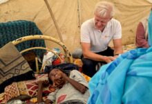 Man sitting beside a lying child inside a tent, surrounded by personal belongings.