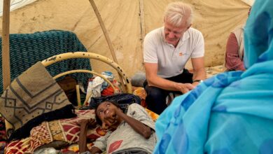 Man sitting beside a lying child inside a tent, surrounded by personal belongings.