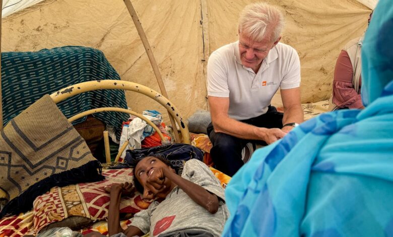 Man sitting beside a lying child inside a tent, surrounded by personal belongings.