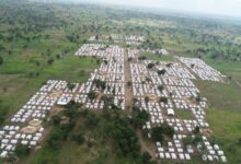 Aerial view of a large refugee camp with rows of white tents scattered across a green landscape.