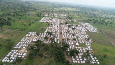 Aerial view of a large refugee camp with rows of white tents scattered across a green landscape.