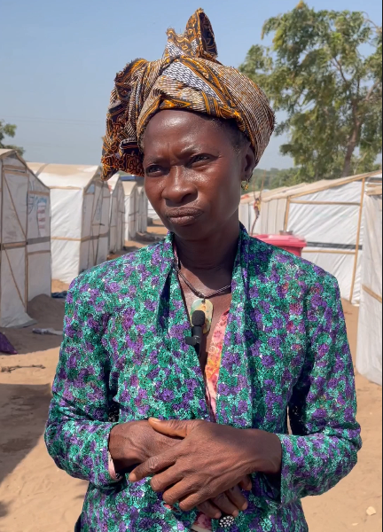 Woman in a colorful blouse and headwrap standing in front of white tents under a clear sky.