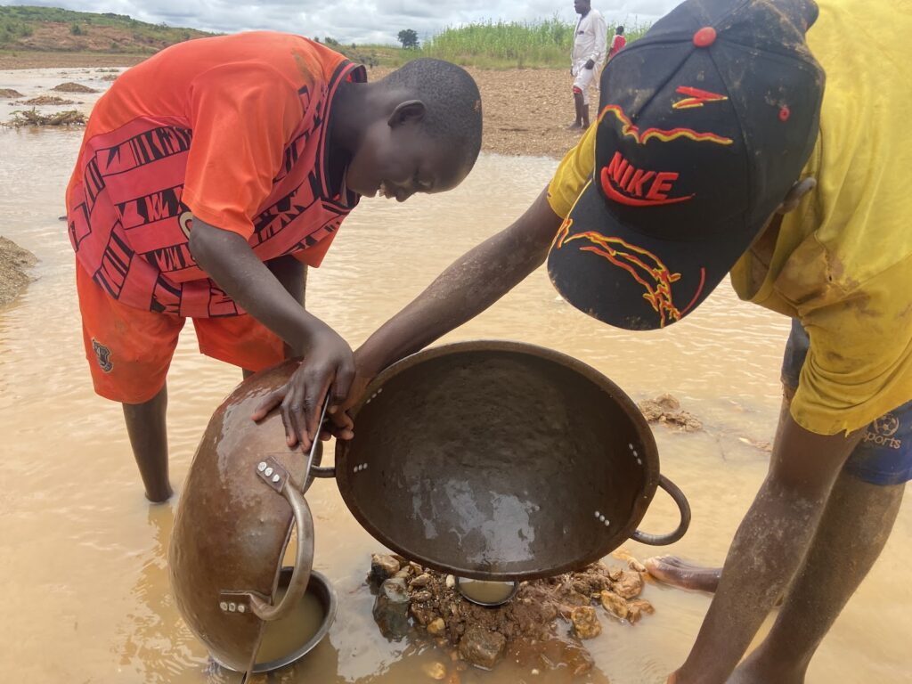 Two individuals sifting material in muddy water, possibly for gold mining, with pans and bowls.