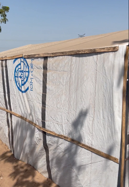 A temporary shelter with white tarp walls displaying the IOM logo under a clear blue sky.