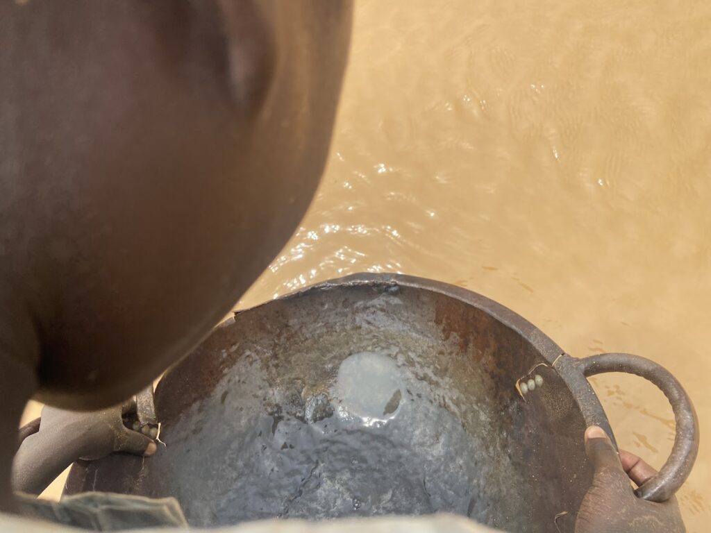 Over-the-shoulder view of a person holding a pan near murky water, possibly panning for minerals.