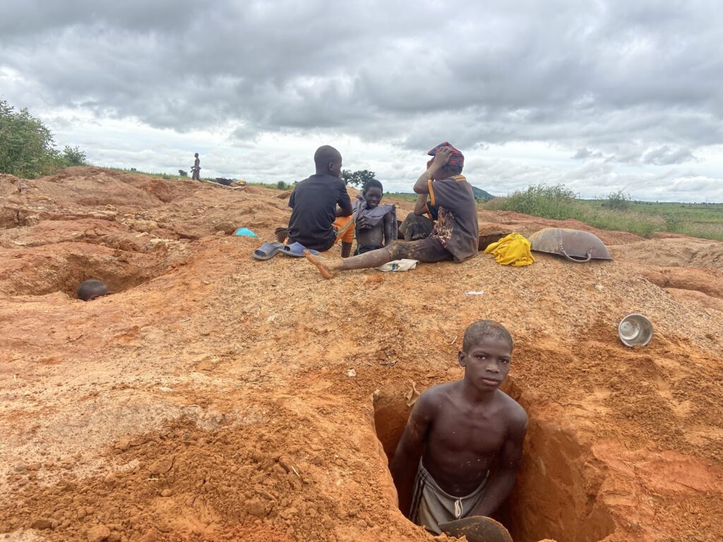 Group of young people on a mining site with red earth, one partially underground, others sitting and resting.