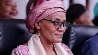 Elderly woman smiling, wearing glasses, pink headscarf, and earrings.