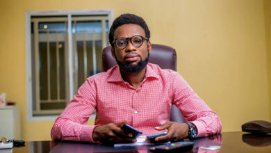 Man in glasses and red checkered shirt sits at desk with smartphone, in office setting.