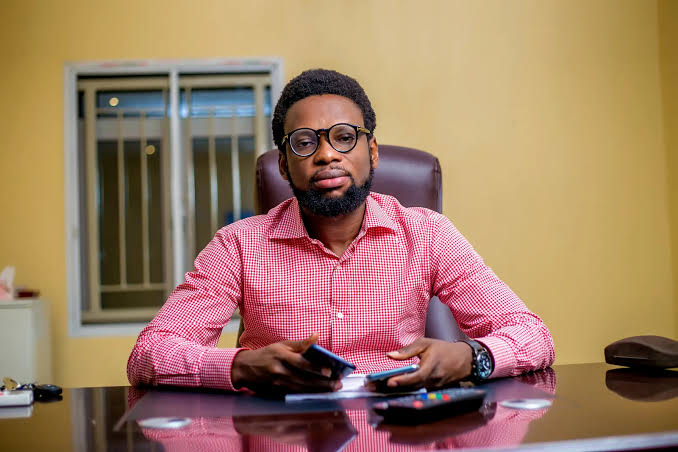 Man in glasses and red checkered shirt sits at desk with smartphone, in office setting.