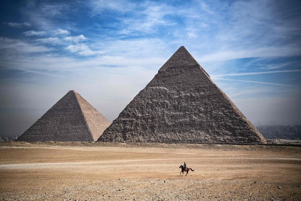 Two ancient Egyptian pyramids under a blue sky with a lone rider on a horse in the foreground.