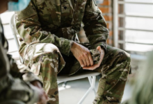 Person in camouflage military uniform sitting with hands clasped, partly cropped view, indoors.