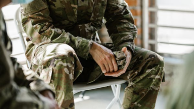 Person in camouflage military uniform sitting with hands clasped, partly cropped view, indoors.