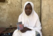 A woman in a white hijab holding a book, sitting by a weathered wall.