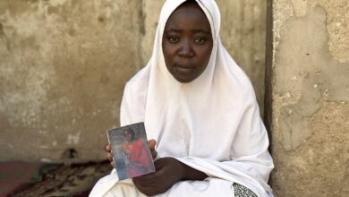 A woman in a white hijab holding a book, sitting by a weathered wall.