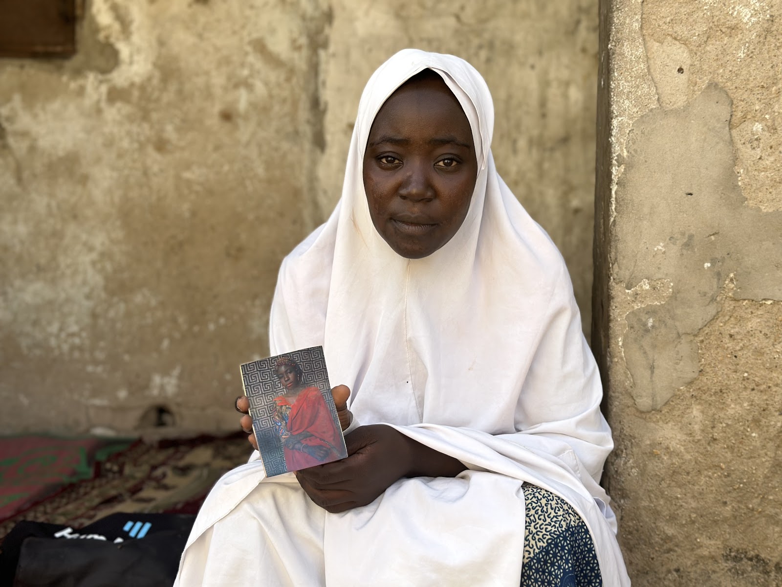 A woman in a white hijab holding a book, sitting by a weathered wall.