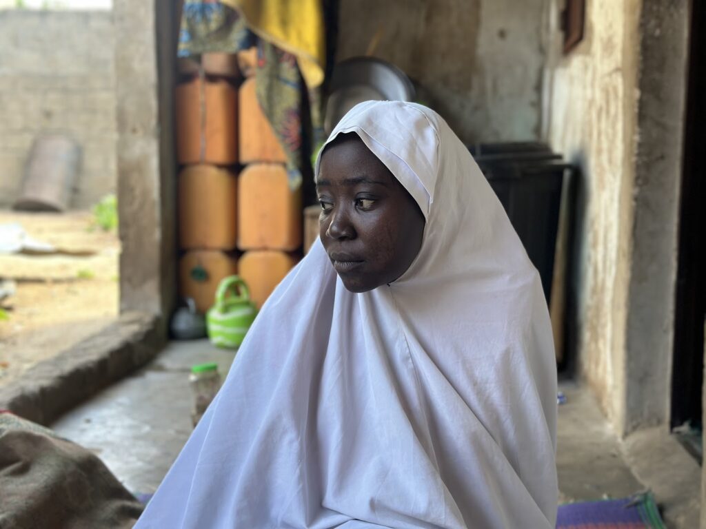 Woman in white hijab sitting pensively in a rustic doorway with buckets in the background.