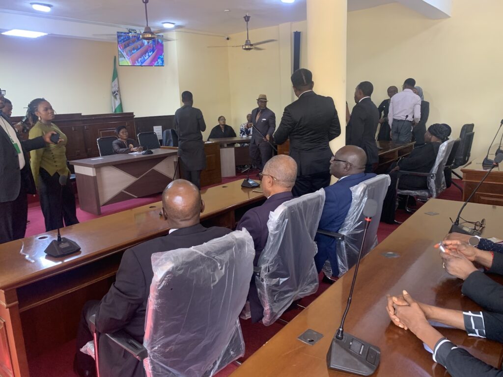 People in a courtroom with a lawyer speaking and others seated, some standing, with chairs covered in plastic.