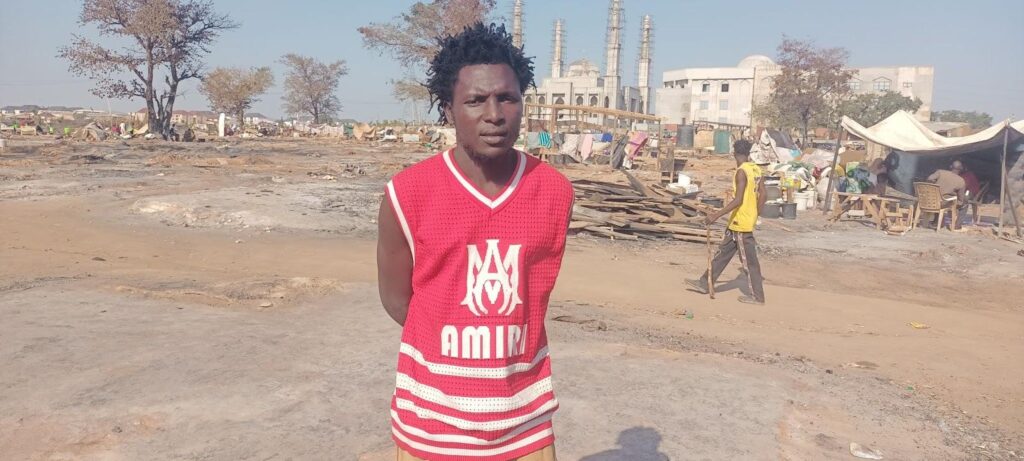 Man in a red basketball jersey standing in front of a makeshift settlement with tents and ongoing activities.