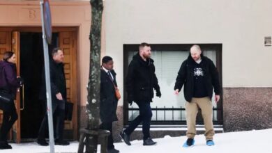 Four individuals walking on a snowy sidewalk by a building, one of them seemingly cautious of slipping.