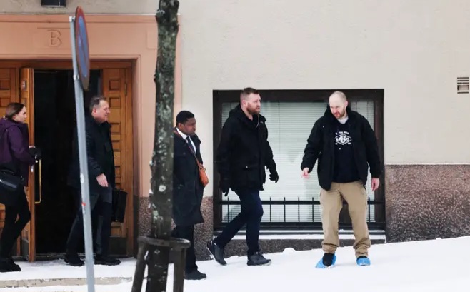 Four individuals walking on a snowy sidewalk by a building, one of them seemingly cautious of slipping.