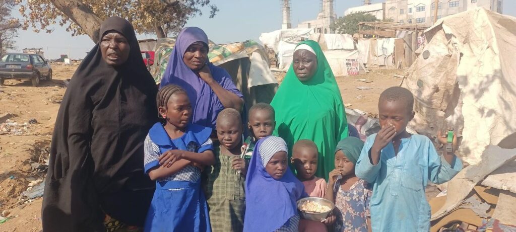 Group of women and children standing in a makeshift camp, with tents and debris around.