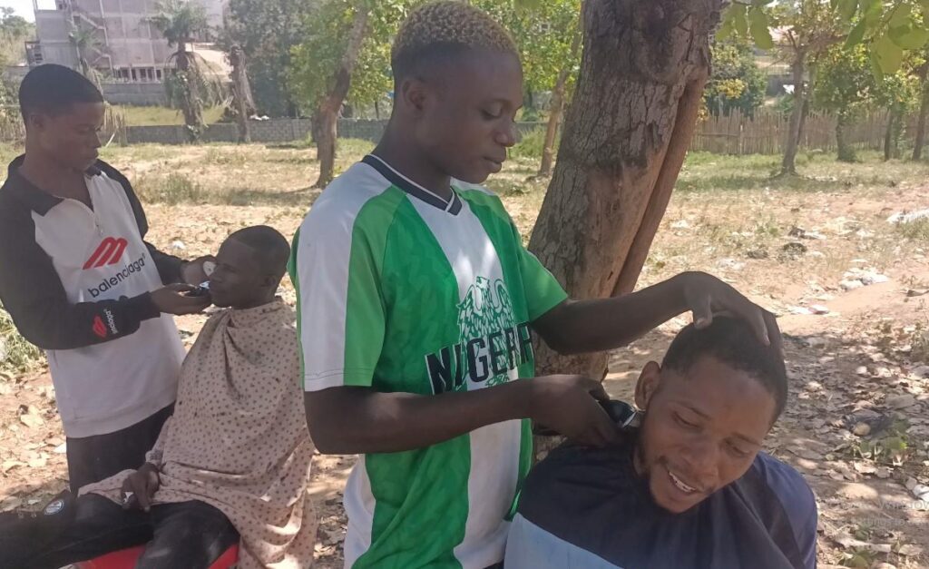 Two individuals giving haircuts under a tree outdoors with one client seated and covered by a cloth.