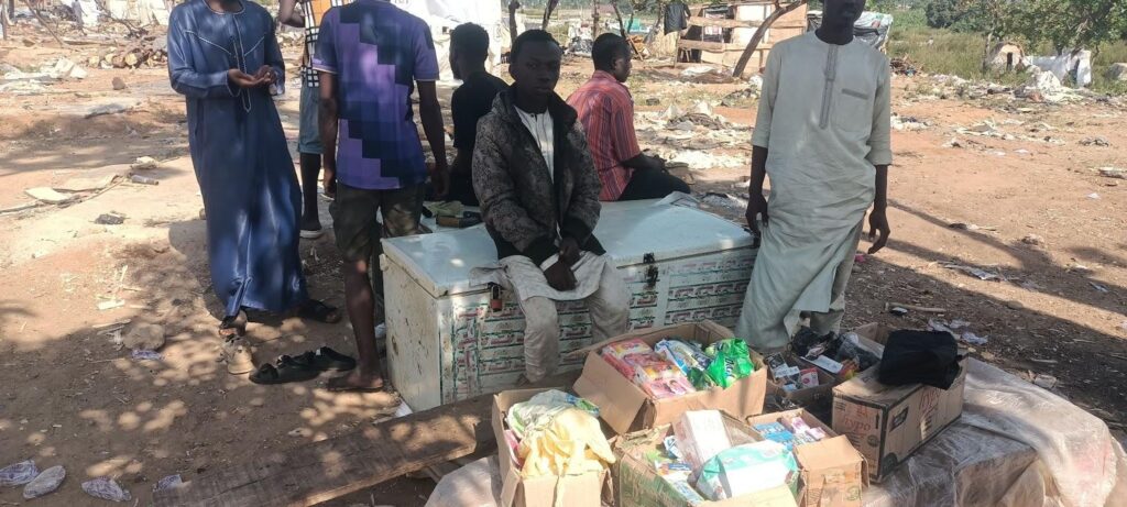 People at an outdoor stall with various goods on display, including a cooler and boxed items, in a rustic setting.