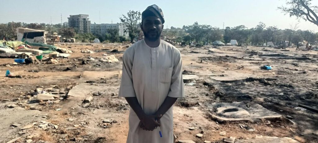 Man in traditional attire stands before a burnt landscape with scattered debris and tents, under a clear sky.