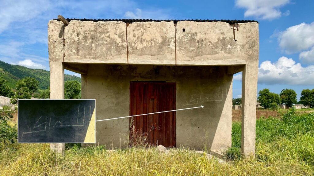 Abandoned concrete structure with a red door, missing roof and walls, surrounded by wild grass, with hills and a clear sky in the background.