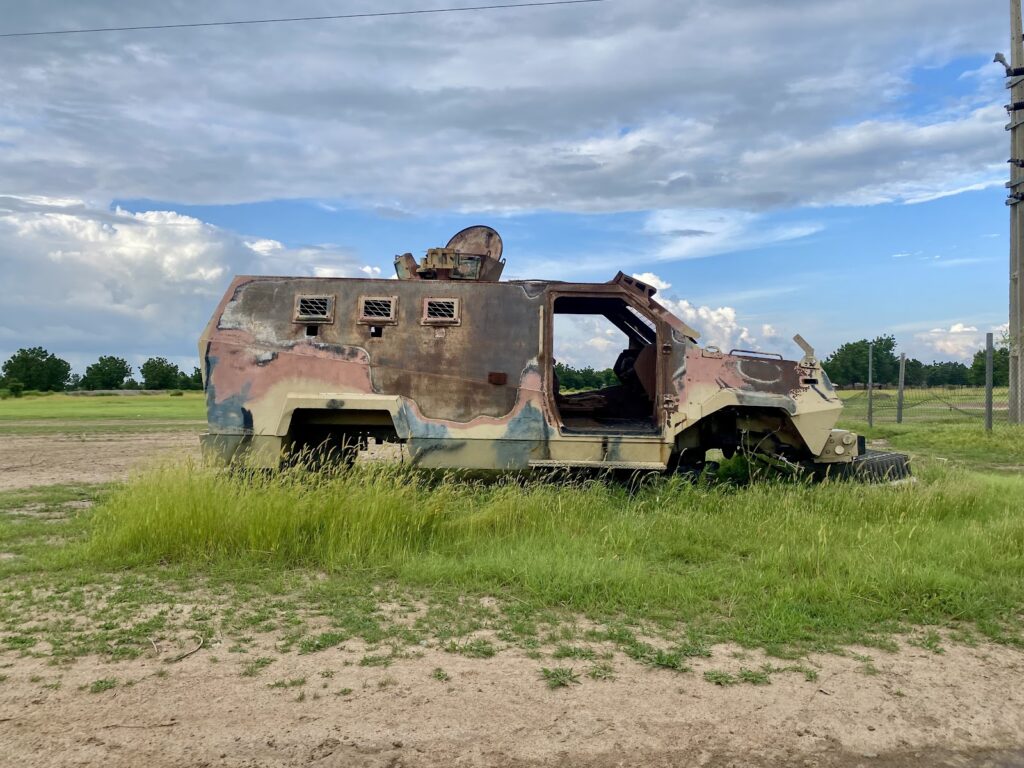 Rusty abandoned armored vehicle in a grassy field with cloudy blue sky.