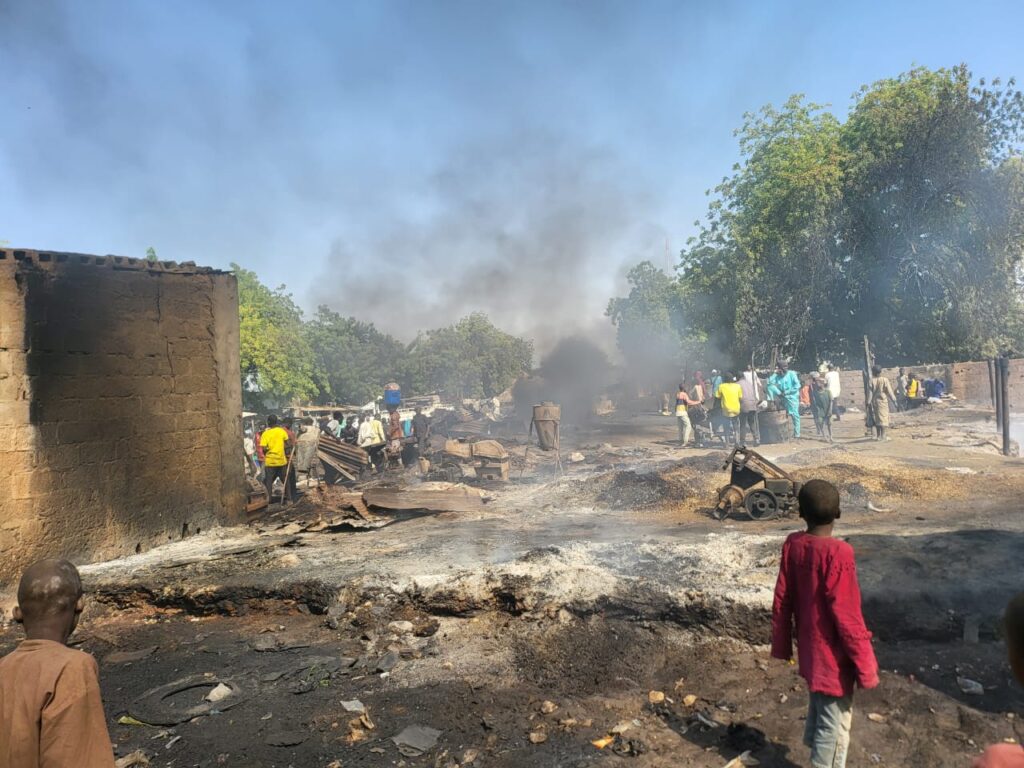 People amidst burnt debris and smoke, with trees in the background.