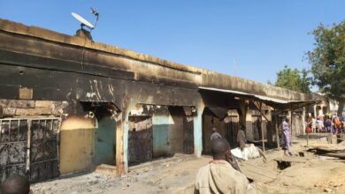 Burnt-down market stalls with onlookers and clear skies, indicating post-disaster scene.