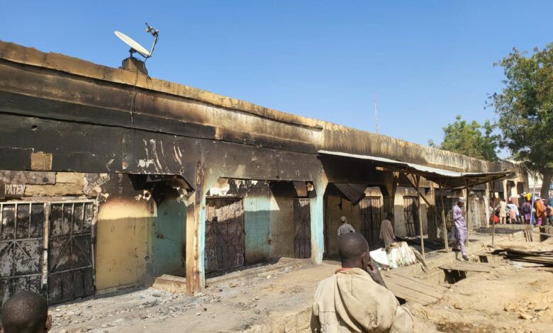 Burnt-down market stalls with onlookers and clear skies, indicating post-disaster scene.