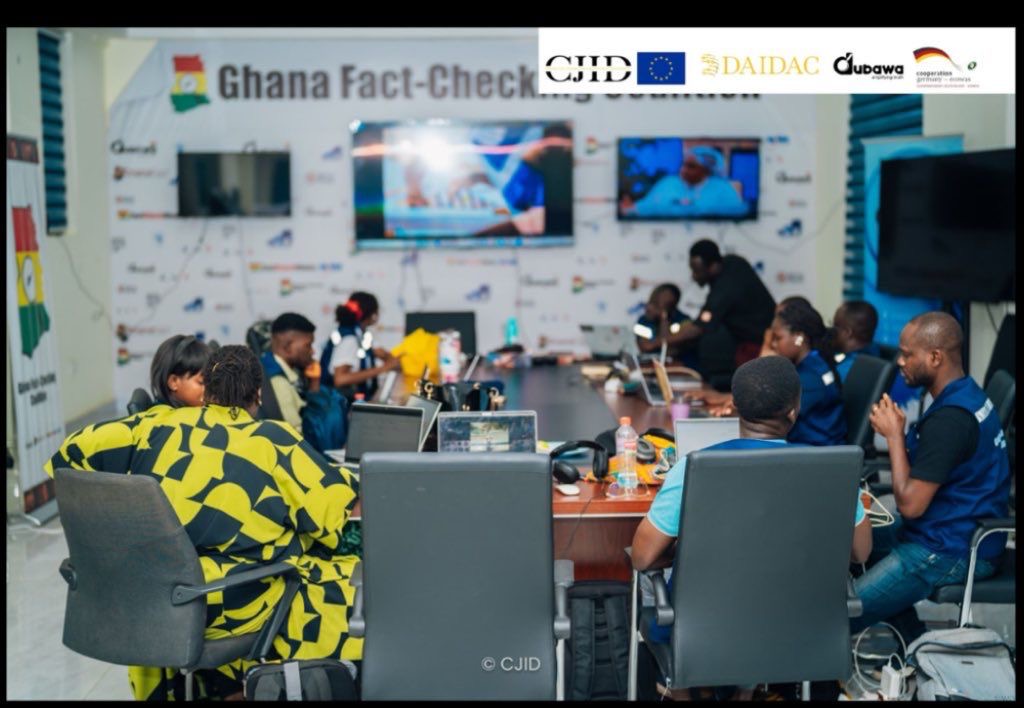 People working in a busy office with computers and monitors, banners for Ghana Fact-Check in the background.