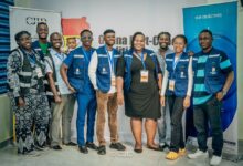 Group of smiling people wearing matching vests and ID badges in front of a "Ghana Fact-Check" banner.