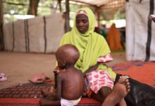 Woman in a green headscarf sits beside a young child on a patterned mat in a sheltered outdoor area.