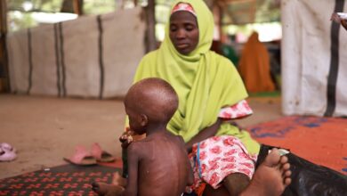 Woman in a green headscarf sits beside a young child on a patterned mat in a sheltered outdoor area.