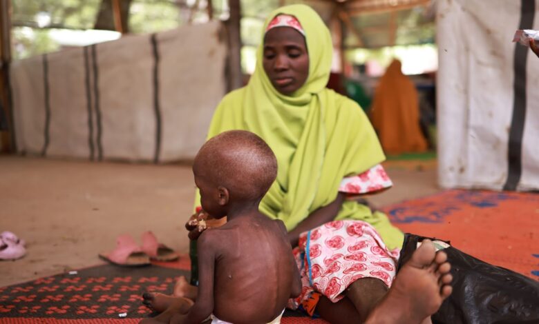 Woman in a green headscarf sits beside a young child on a patterned mat in a sheltered outdoor area.