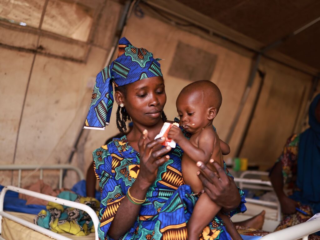 Woman in colorful attire holding a young child and a medicine bottle in a clinic setting.