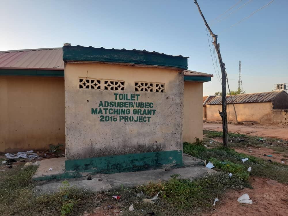 A small public toilet building with a sign indicating it's part of a 2016 project, surrounded by some litter.