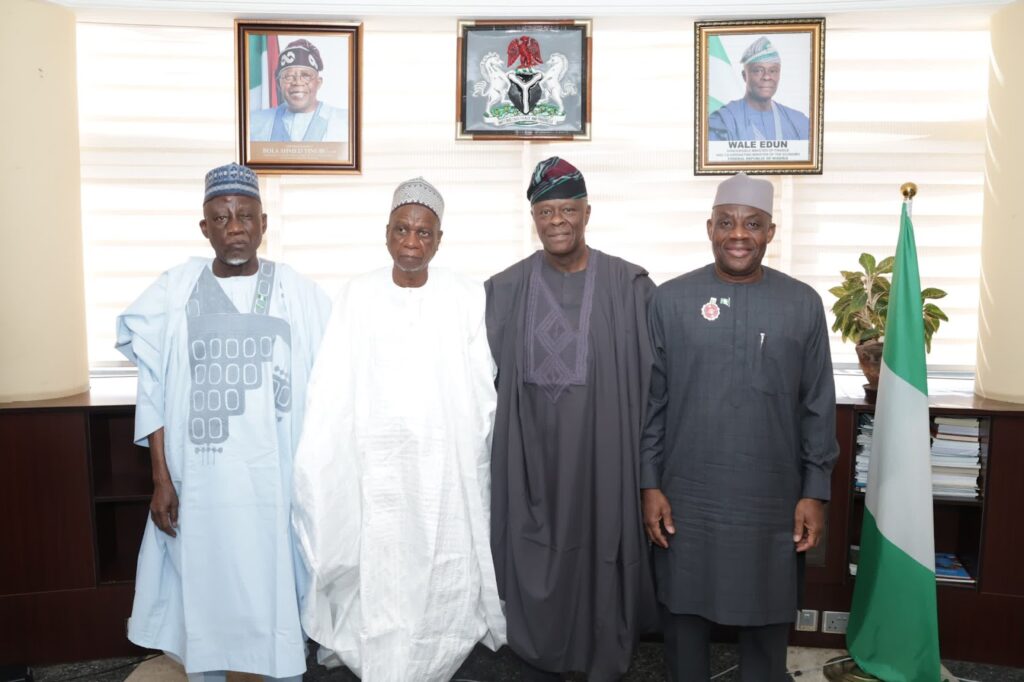 Four men in traditional and formal attire standing in an office with framed portraits and a Nigerian flag.