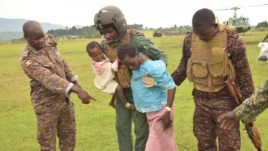 Soldiers assisting a woman and child in a grassy field with a helicopter in the background.