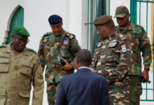 Four military officers in camo uniforms talking with a man in a suit, outdoors near a building.