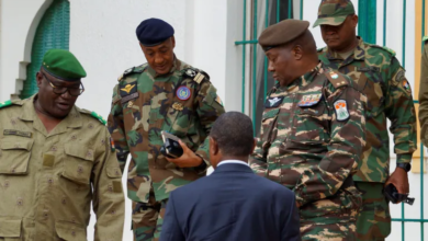 Four military officers in camo uniforms talking with a man in a suit, outdoors near a building.