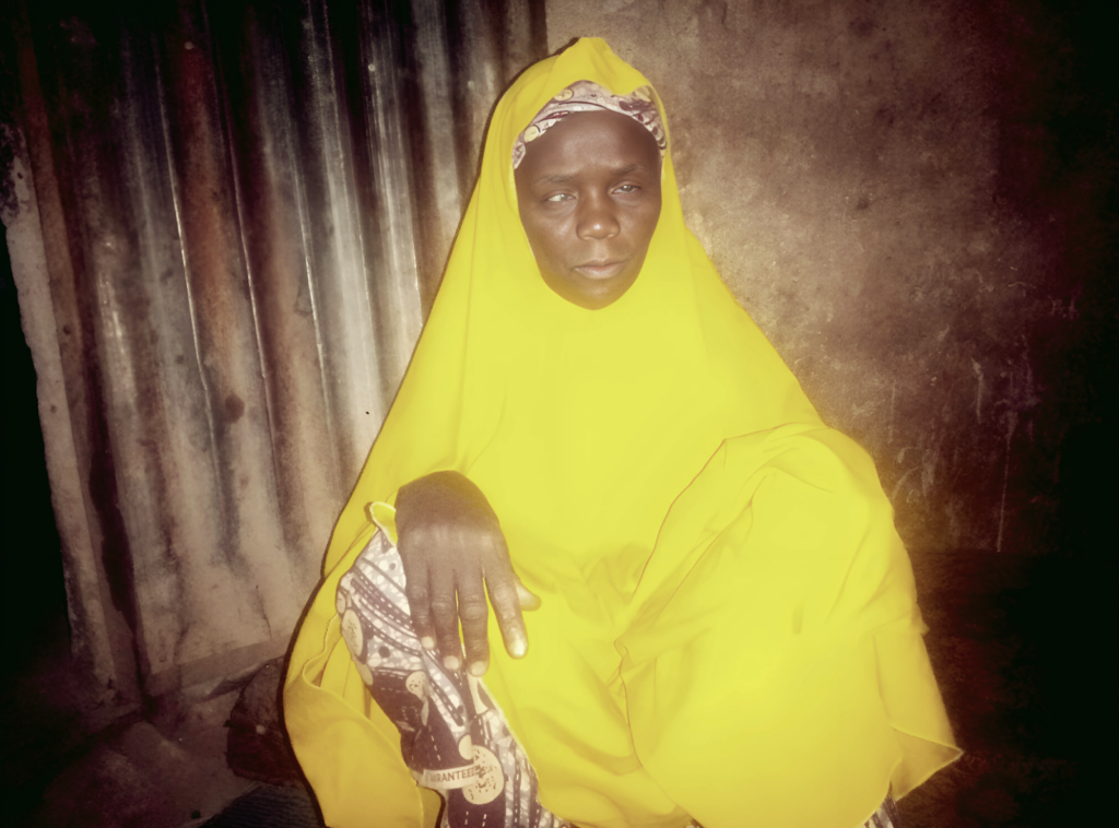 Woman in a bright yellow garment sitting in front of a corrugated metal background.